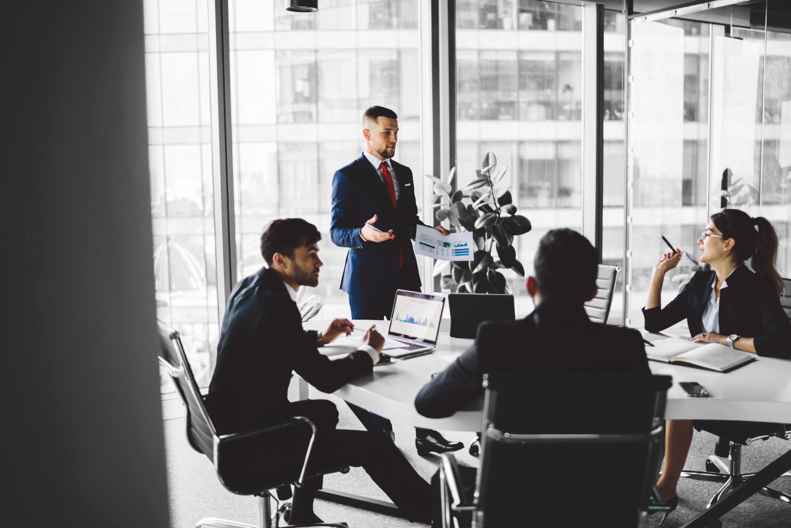 Confident successful businessman in formal black suit standing next to colleagues and analyzing financial report during conference in modern workspace