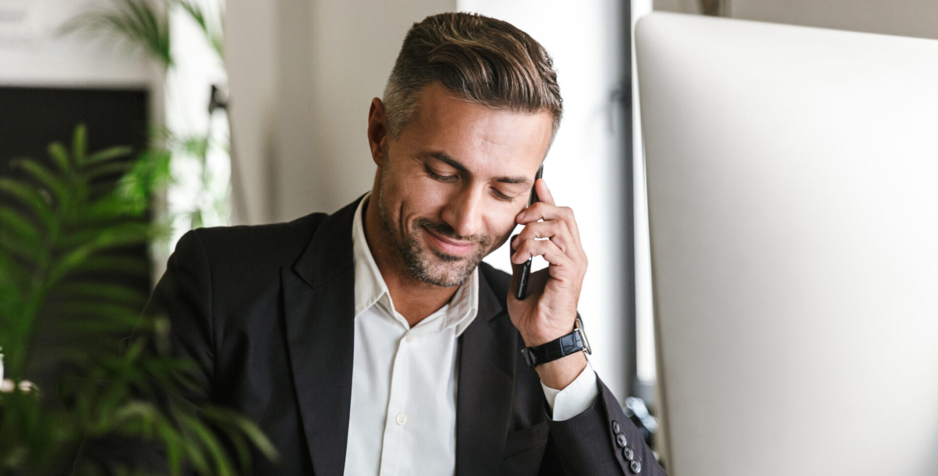 Image of handsome businessman talking on cell phone about remittance while working on computer in office