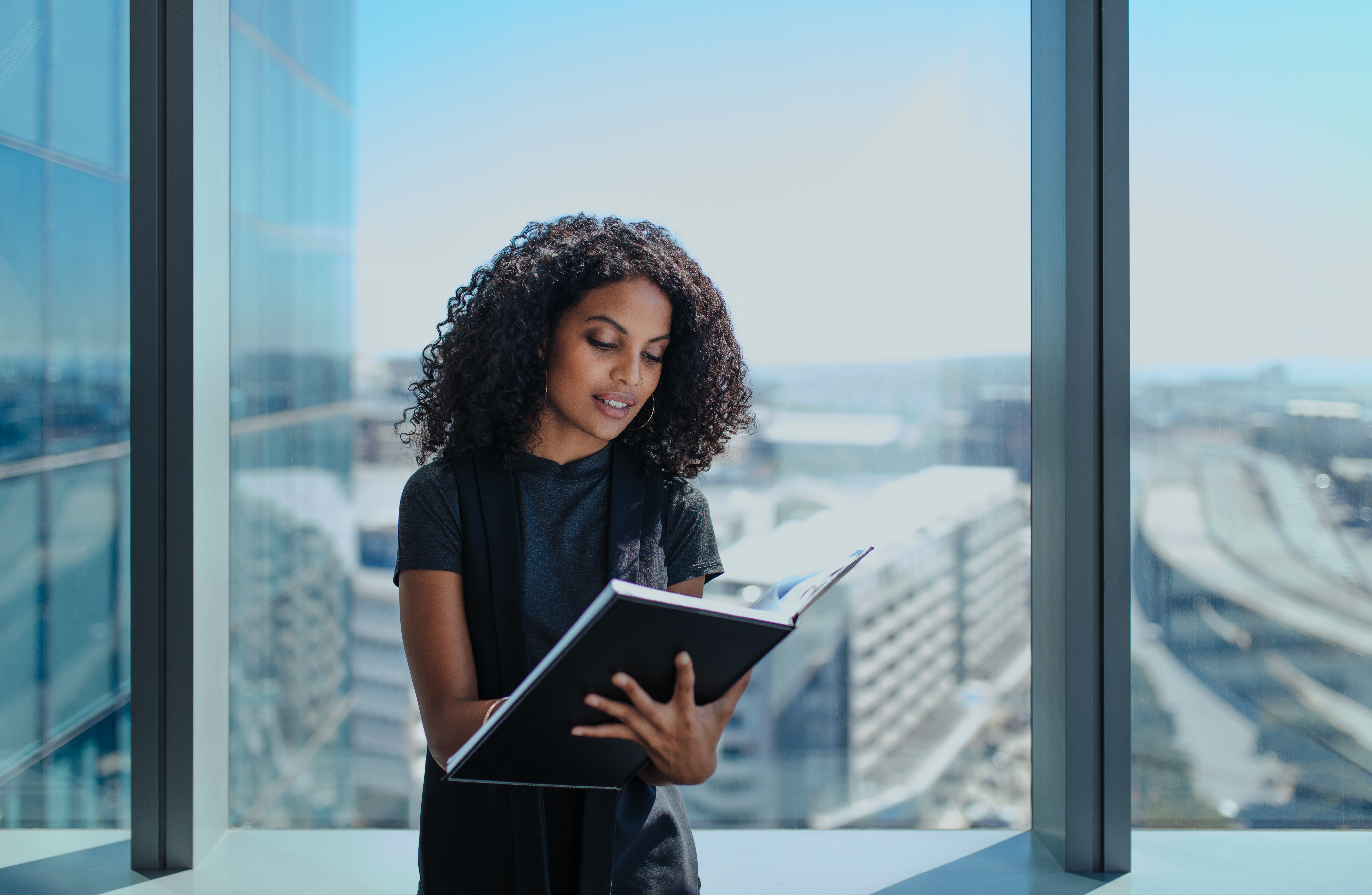 Businesswoman writing in a book standing near window in office. Businesswoman standing in her office in a highrise building overlooking the cityscape.