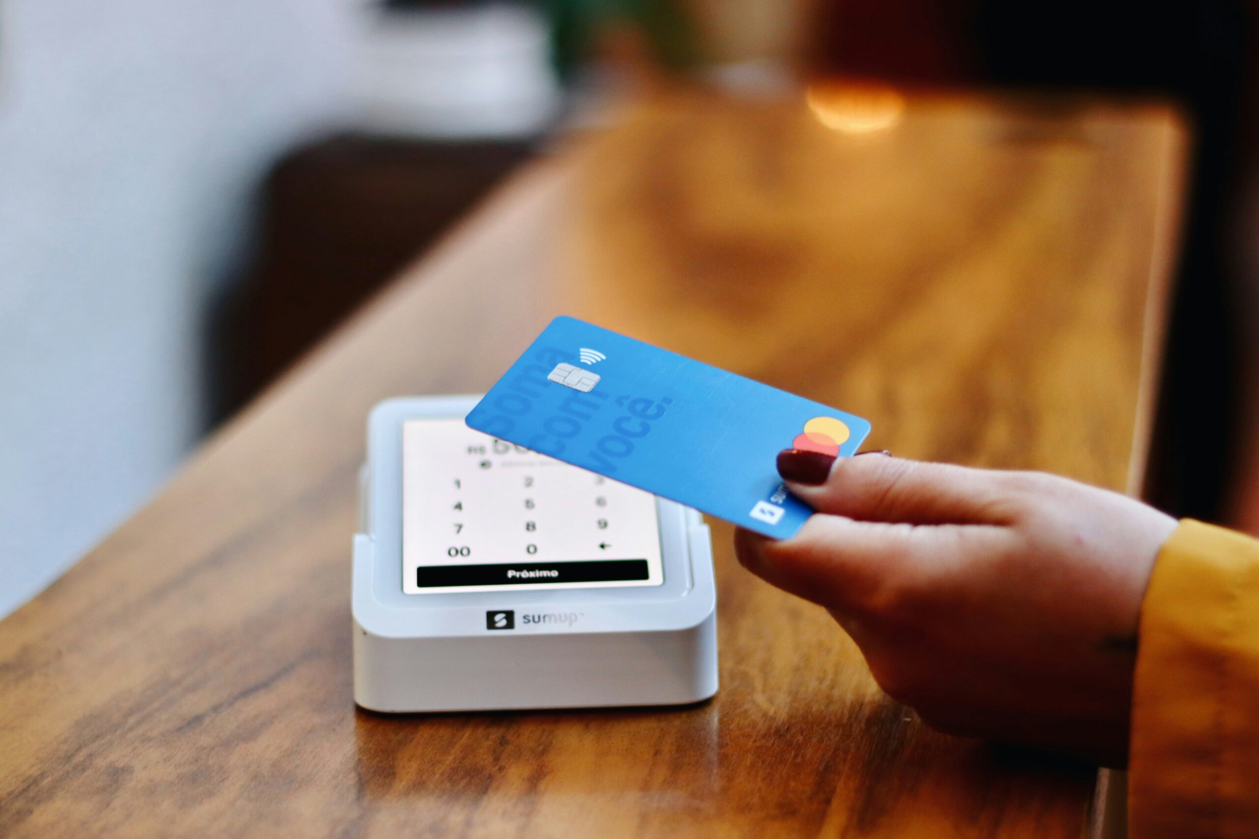 A close-up image shows a payment terminal being used. In the foreground, a blue contactless payment card is being held just above the terminal, moments before or during a tap-to-pay action.