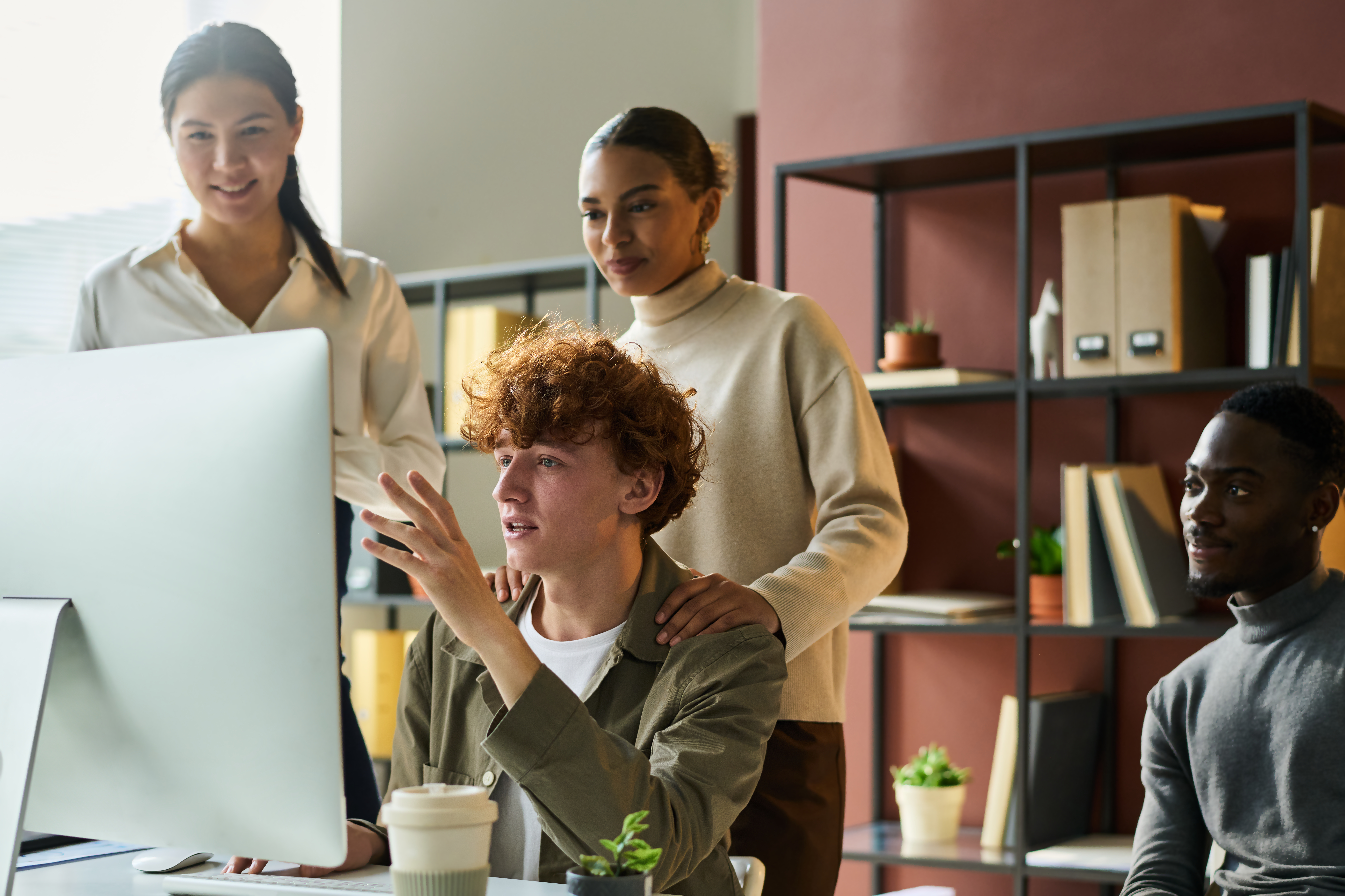 A group of young professionals gathers around a desktop monitor, engaged in a collaborative discussion. Their focused expressions and team dynamic suggest they are reviewing AI-powered insights to improve financial close workflows. The setting, with bookshelves and plants, reflects a modern and organized workspace.
