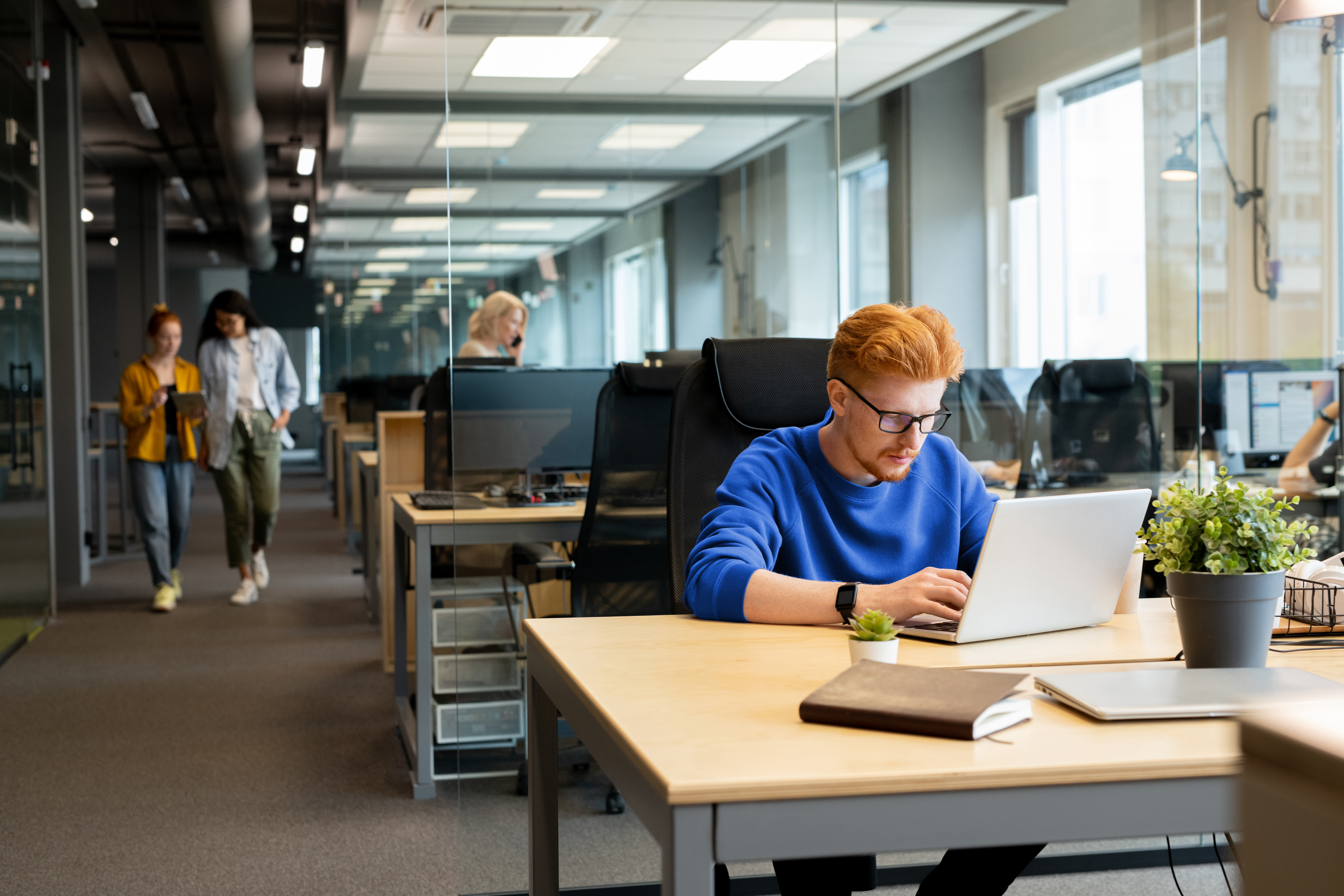 A focused man in a blue sweater works on a laptop in a modern, open-plan office. He appears to be leveraging AI software to enhance the financial close process. The workspace features glass partitions, greenery, and a clean, tech-driven atmosphere, with colleagues collaborating in the background.