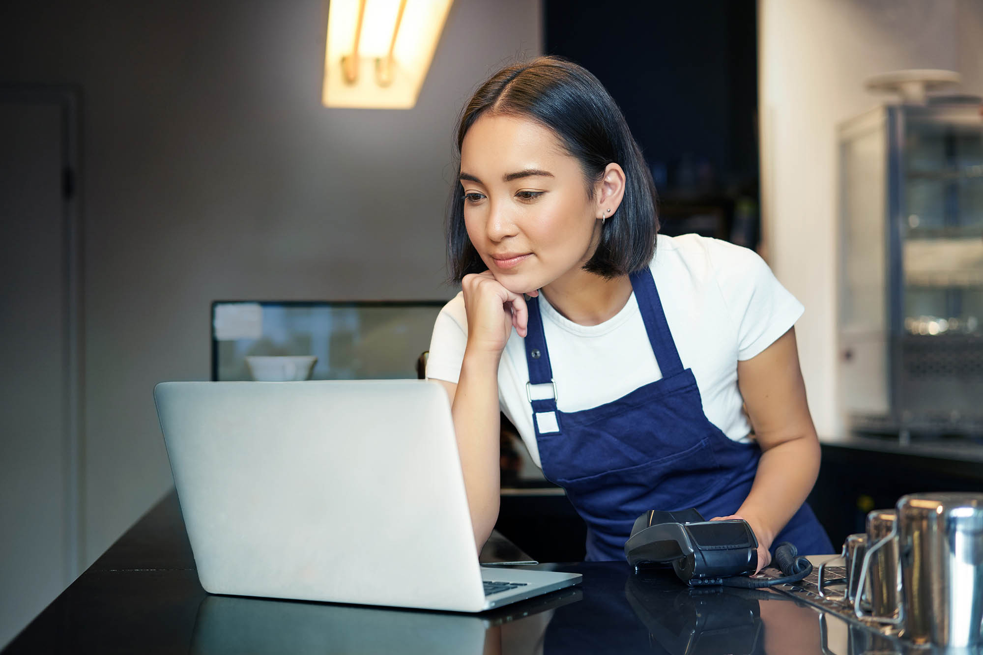 Restaurant employee reviewing account reconciliation tasks on laptop at counter with payment terminal